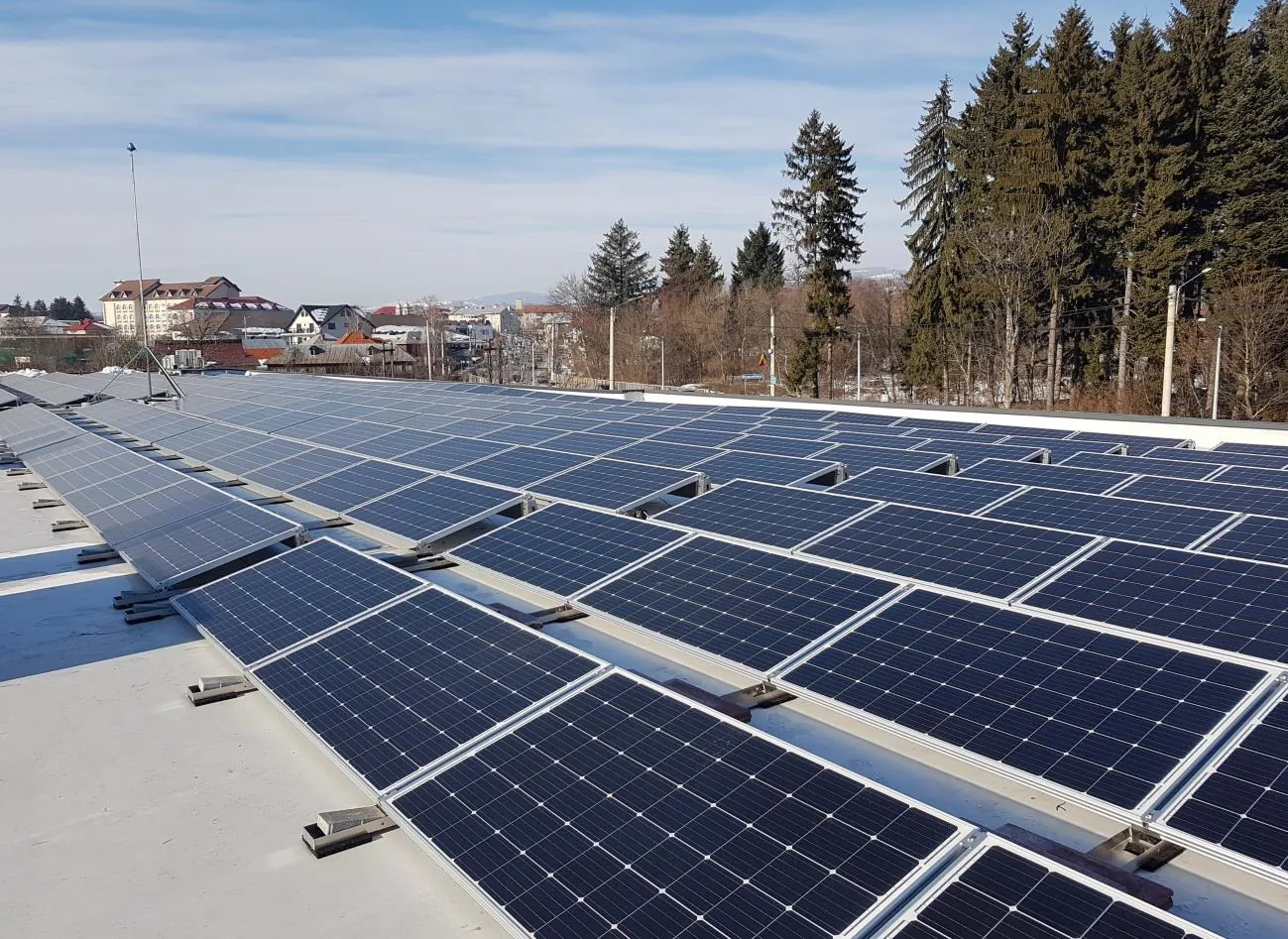 Panouri fotovoltaice montate pe acoperișul plat al unei hale, cu localitatea în fundal